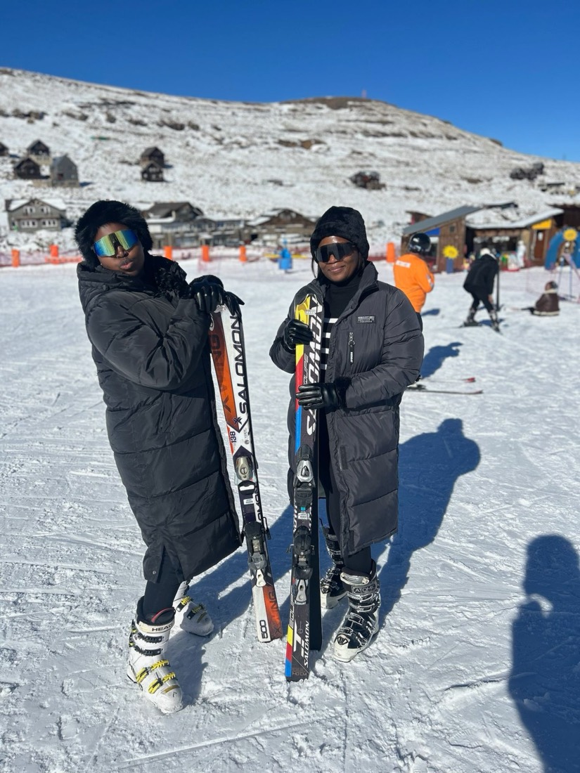 Two friends in long black puffer coats holding skis on the slope at Afriski, Lesotho